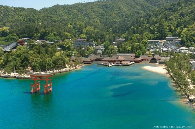 Vista aérea de la bahía de Miyajima con el santuario en la orilla, el torii en aguas turquesa y colinas boscosas detrás